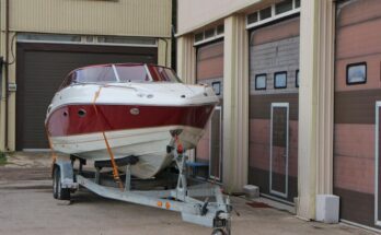 A large white boat with red accents on top of a silver trailer next to a row of closed storage locker doors.