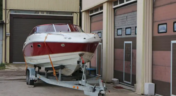 A large white boat with red accents on top of a silver trailer next to a row of closed storage locker doors.