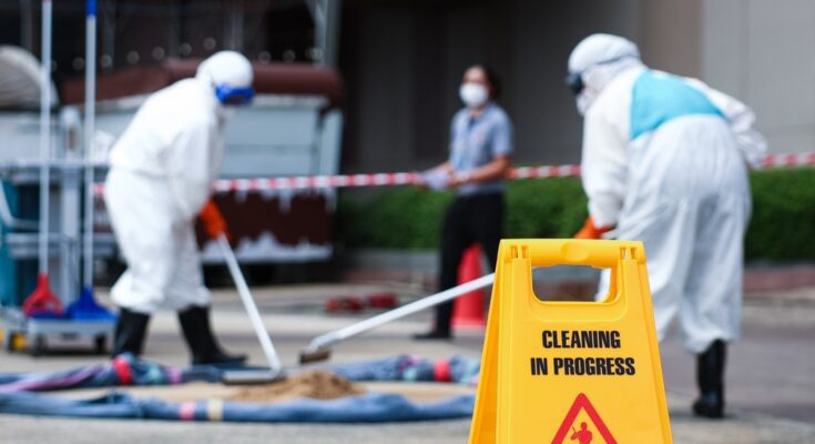 A close-up of a yellow hazard sign with two workers behind it in white hazmat suits. They're cleaning a spill.