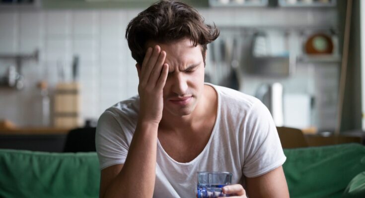 A man wearing a white shirt sits on a green couch holding a glass of water while rubbing his face with his hand.