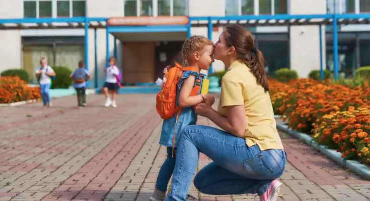 A woman wearing a yellow shirt and jeans kisses a child on the forehead. The young child wears an orange backpack.