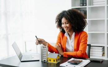 A woman wearing an orange coat smiles while sitting at a desk with a laptop, models, and a calculator on top of paper.