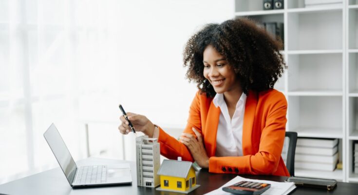 A woman wearing an orange coat smiles while sitting at a desk with a laptop, models, and a calculator on top of paper.