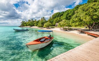 Two motorized boats move through turquoise water near a sandy beach and wooden dock, with a blue sky above.