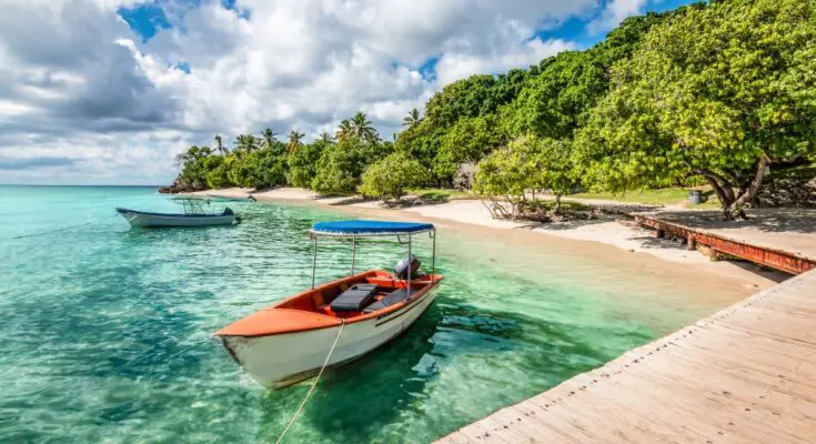 Two motorized boats move through turquoise water near a sandy beach and wooden dock, with a blue sky above.