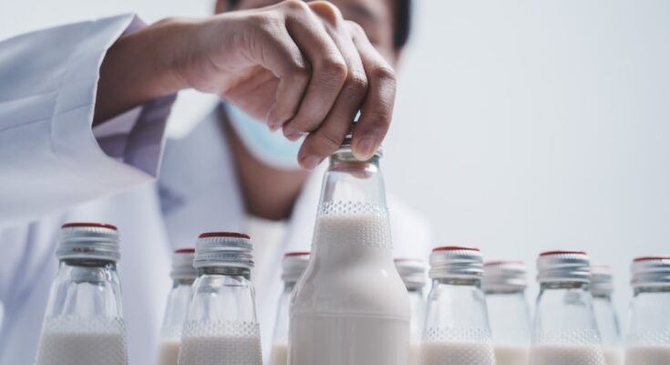 A person wearing a mask and a white lab coat standing in front of many milk bottles and inspecting the cap on one.