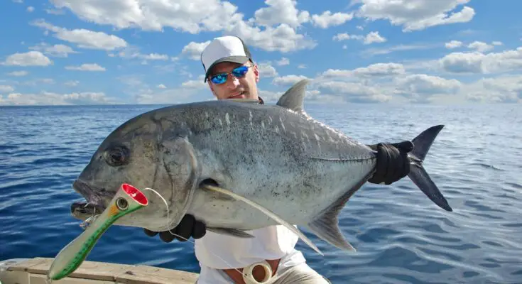 A happy angler sits on the edge of a charter boat at sea as he holds up a large fish. He's wearing blue sunglasses.