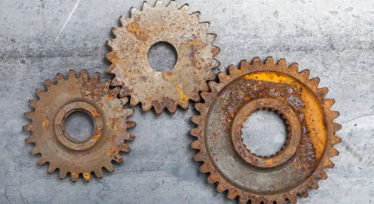 Three rusted metal gears of different sizes sit interlocked with each other on a gray concrete floor.