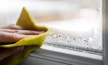 A close-up of a hand holding a sponge, wiping off water condensation from the plastic window glass in the room.