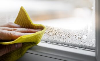 A close-up of a hand holding a sponge, wiping off water condensation from the plastic window glass in the room.