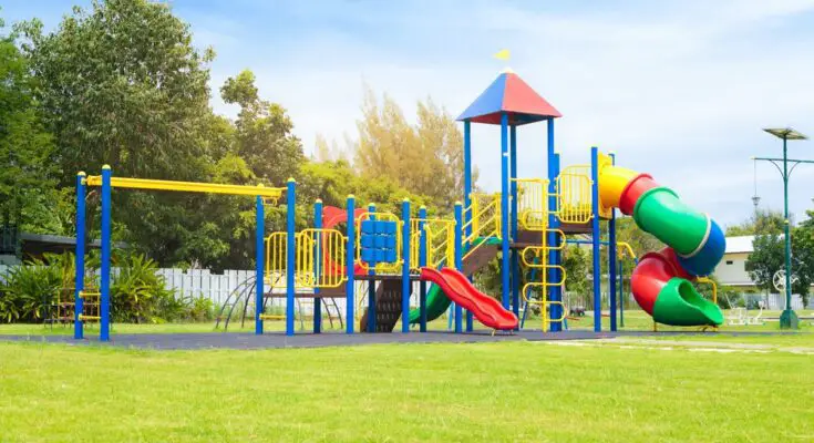 A colorful playground contains a twisty slide, monkey bars, stairs, and two straight slides. It sits on gray surfacing.