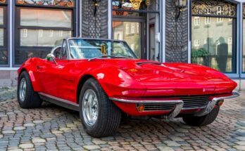 A red classic car is parked in front of a historic building on a cobblestone road with a mini flag on the rearview mirror.
