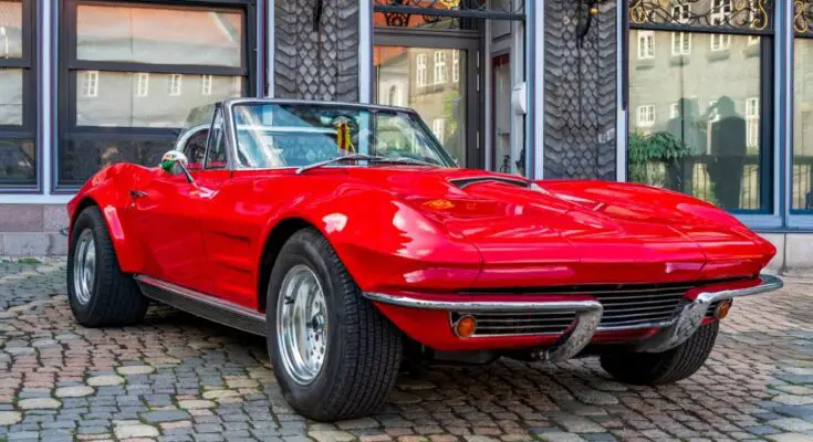 A red classic car is parked in front of a historic building on a cobblestone road with a mini flag on the rearview mirror.