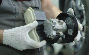 A person wearing gloves holds a hydraulic pump in their hands. A car tire is visible in the background.