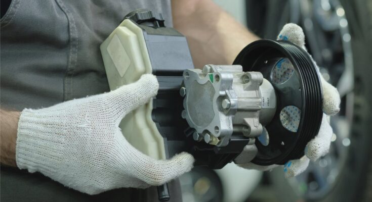 A person wearing gloves holds a hydraulic pump in their hands. A car tire is visible in the background.