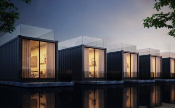 A row of shipping container homes are sitting along a body of water on a dim evening. The windows are lit, showing the interiors.