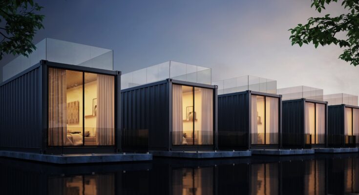 A row of shipping container homes are sitting along a body of water on a dim evening. The windows are lit, showing the interiors.