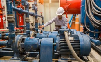 A person wearing a white hard hat holds a table while inspecting three large, blue industrial motors.