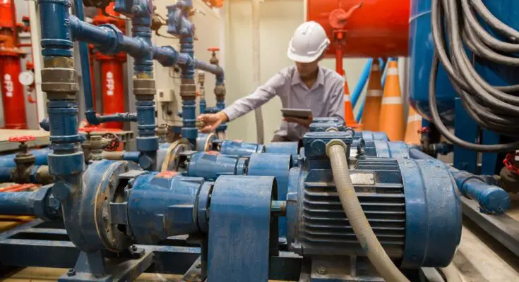 A person wearing a white hard hat holds a table while inspecting three large, blue industrial motors.