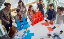 Two groups of coworkers compete to build a house of cards with paper. One team uses red paper; the other uses blue.