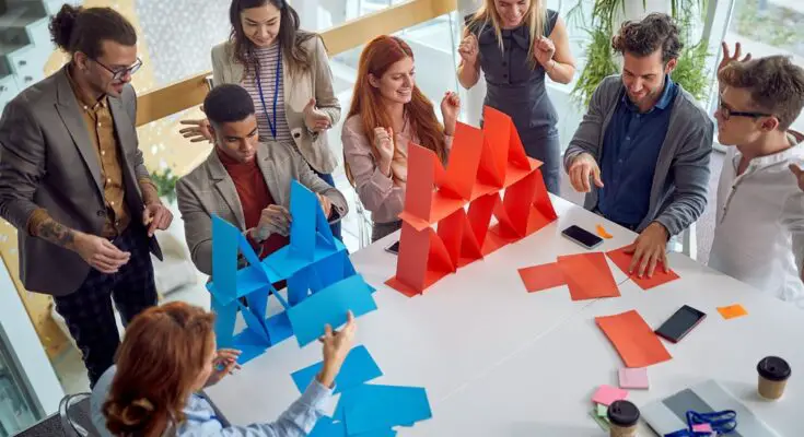 Two groups of coworkers compete to build a house of cards with paper. One team uses red paper; the other uses blue.