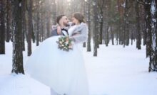 A groom in a gray suit holds his bride in a snowy forest; she wears a voluminous gown, fur jacket, and carries a bouquet.