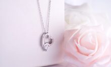 A silver necklace with a heart-shaped pendant and a diamond hangs over a jewelry box. A pink rose is beside the box.