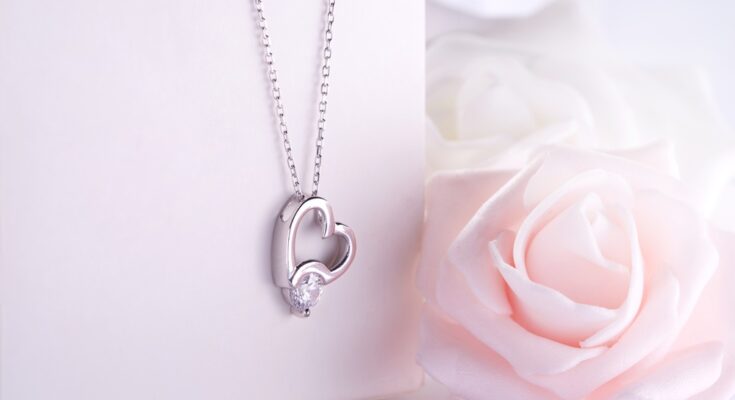 A silver necklace with a heart-shaped pendant and a diamond hangs over a jewelry box. A pink rose is beside the box.