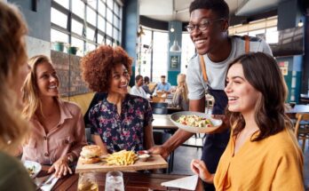 A smiling waiter holding a tray with a hamburger and fries and a bowl of salad over a table surrounded by smiling customers.