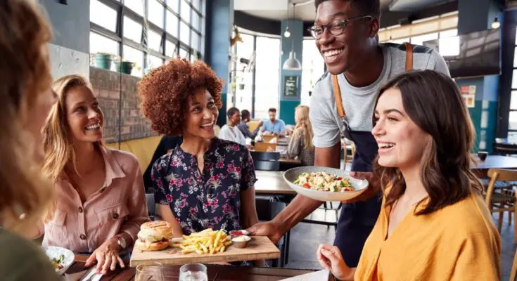 A smiling waiter holding a tray with a hamburger and fries and a bowl of salad over a table surrounded by smiling customers.