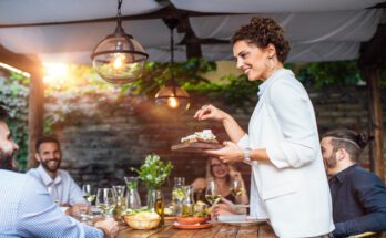 A woman is hosting a dinner party and is serving food to her friends while pointing to the dish she's serving.