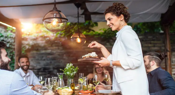 A woman is hosting a dinner party and is serving food to her friends while pointing to the dish she's serving.