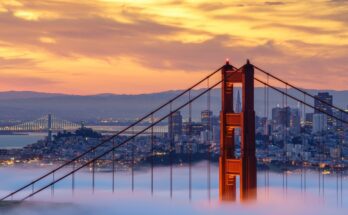 A red tower of the Golden Gate Bridge rises above low-lying fog. The city skyline and mountains are in the background.