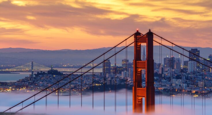 A red tower of the Golden Gate Bridge rises above low-lying fog. The city skyline and mountains are in the background.