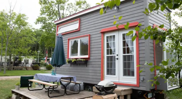 A close-up of a modern tiny home. The home is gray and red with a wooden outdoor seating area and grill.