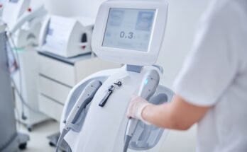 A person wearing personal protective gear grabs a part of a medical device in a room filled with other equipment.