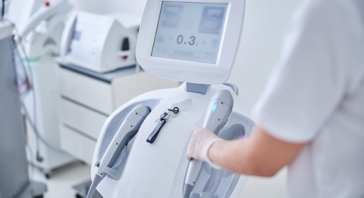 A person wearing personal protective gear grabs a part of a medical device in a room filled with other equipment.