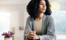 A woman sitting at a table has her fingers laced together and her hands resting on a closed book. She looks to her left.
