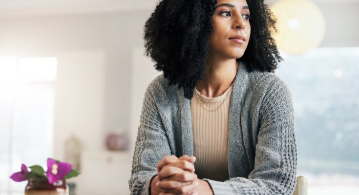 A woman sitting at a table has her fingers laced together and her hands resting on a closed book. She looks to her left.