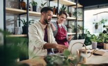 Two shop assistants are talking, wearing aprons, and working on their laptops in a small potted plant business.