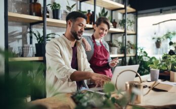 Two shop assistants are talking, wearing aprons, and working on their laptops in a small potted plant business.