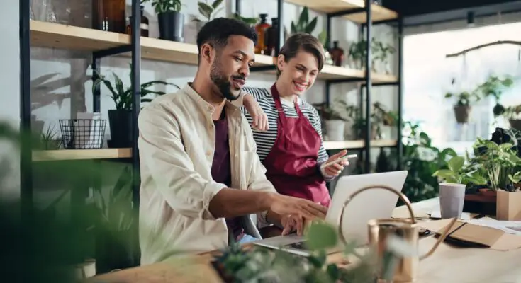 Two shop assistants are talking, wearing aprons, and working on their laptops in a small potted plant business.