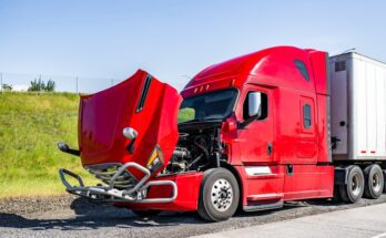 A red semi-truck parked next to a freeway near a grassy hill in the middle of the day. The vehicle’s hood is propped open.