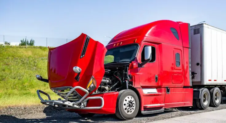 A red semi-truck parked next to a freeway near a grassy hill in the middle of the day. The vehicle’s hood is propped open.