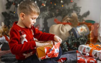 A young boy sits in front of the Christmas tree, wearing a red Christmas sweater and opening wrapped presents.