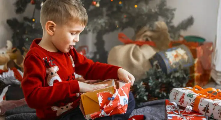 A young boy sits in front of the Christmas tree, wearing a red Christmas sweater and opening wrapped presents.