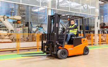 A man in a hi-vis yellow safety vest drives an orange forklift around a warehouse. There's an orange fence behind him.