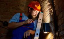 A man with a beard wearing a hard hat and overalls holds a screwdriver up to an outdoor lighting fixture on a brick wall.