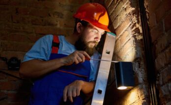 A man with a beard wearing a hard hat and overalls holds a screwdriver up to an outdoor lighting fixture on a brick wall.