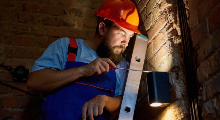 A man with a beard wearing a hard hat and overalls holds a screwdriver up to an outdoor lighting fixture on a brick wall.